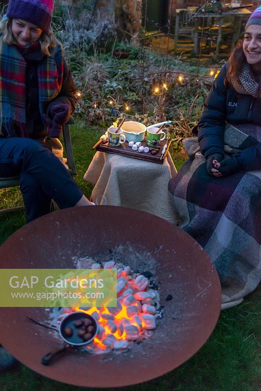 Woman wrapped in warm clothes, cooking chestnuts over a fire in a Corten Steel fire pit on a winter evening