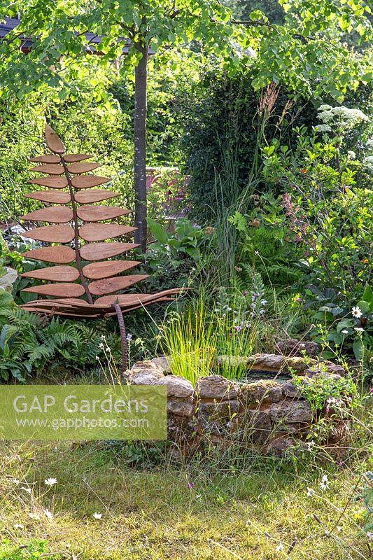 Bespoke leaf-shaped chair in wild garden with naturalistic planting and a central circular stone water feature. Calm Admidst Chaos, Hampton Court Flower Festival, 2019.