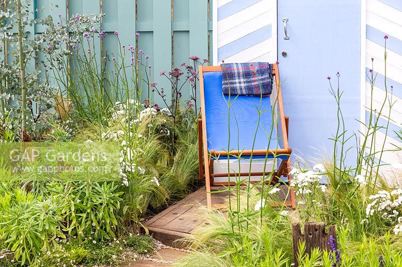 Blue and white painted beach huts with deck chairs on decking with Achillea 'Wonderful Wampee', Nepeta racemosa 'Walker's Low', Verbena bonariensis, Stipa tenuissima. By The Sea, RHS Hampton Court Palace Flower Show, 2017. Design: James Callicott, Sponsors: Southend.
