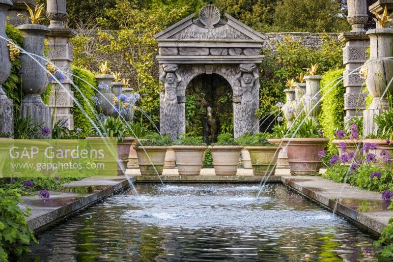 The Collector Earl's Garden at Arundel Castle, West Sussex in May. Designed by Isabel and Julian Bannermann. Green oak urns, pillars and a grotto guarded by sea gods frame water representing the alegorical River Arun. Gilded lions and agaves adorn the urns, and planting includes alliums, agapanthus and Alchemilla mollis.
