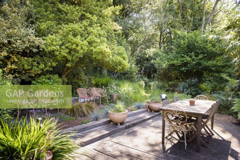 Simple decked dining area surrounded by dense trees and shrubs in a cottage garden in June. Four contemporary orange metal seats are arranged in a row to one side with planting including Carex testacea and blue festuca.