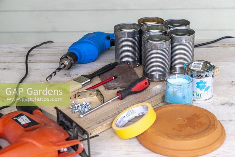 Tin cans, drill, jigsaw, wooden board, screwdriver, screws, brackets, pencil, ruler, paintbrush, paint, sandpaper, masking tape and terra cotta dish laid out on a wooden surface