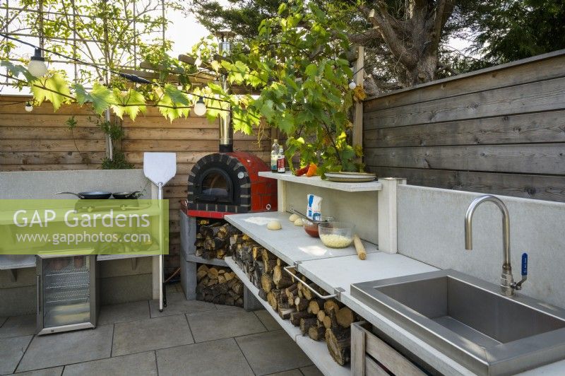 Outdoor kitchen with red pizza oven, metal sink, hob and wood storage in small modern family garden