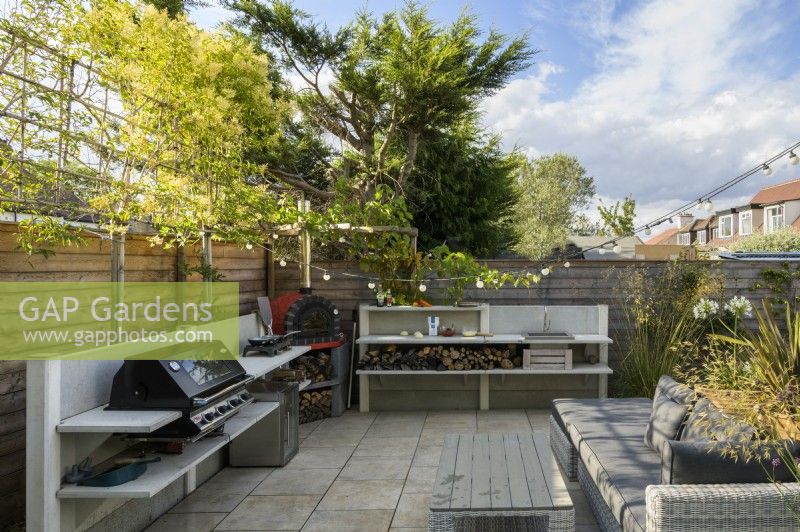 Outdoor kitchen with red pizza oven, hob and barbecue, with built-in wood storage and pleached trees for privacy in small modern family garden