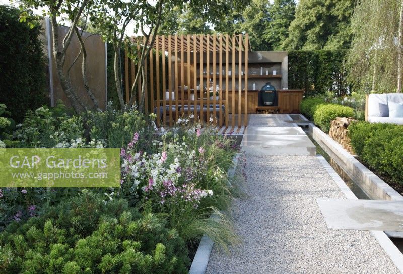 View of The Viking Friluftsliv Garden where a long path leads to an oudoor kitchen and pergola, it is bordered on the left with mixed planting including Pinus mugo 'Mops', and on the right, a long and narrow rill - Designer: Will Williams -Sponsor: Viking
