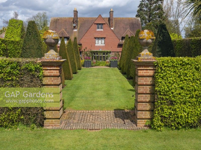 Brickwork pillars and paving marking entrance through hedge to grass path. View of Kings Walk, East Ruston Old Vicarage Gardens, Norfolk, UK.