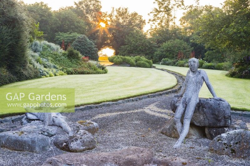 Figures of two naked males in gravel area with view to rill and lawn at sunrise. Sculptures by Jane Hazelwood. July