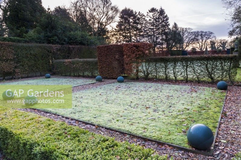 Diagonal view across the frosty lawns as the sun rises. Painted balls form man-made structure alongside the clipped hornbeam and box hedges.