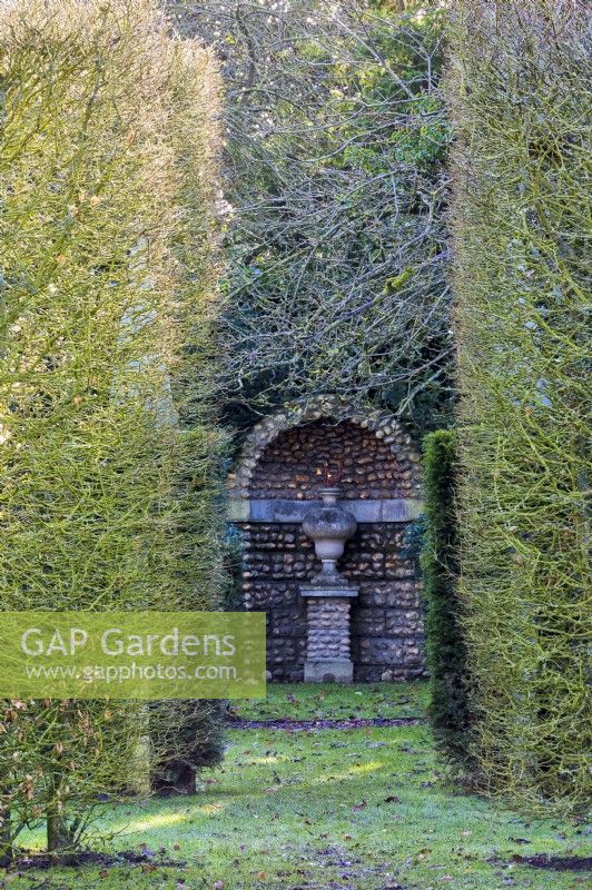 A classical stone urn with a twist in a flint grotto forms the focal point of a vista down the garden. Hornbeam and yew hedges frame.