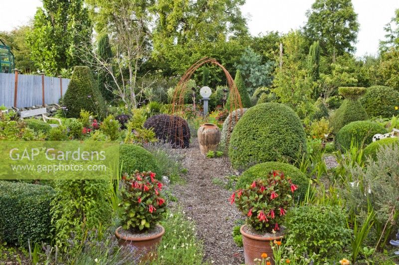 Informal garden with Fuchsia 'Thalia' in pots, clipped topiary mixed, view along gravel path to urn framed by rusty arch.