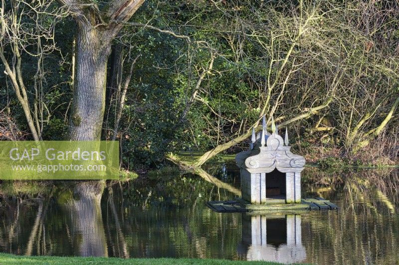 Floating duck shelter: the ornate mini temple with its spiking obelisks dominates the simple woodland pond in shafts of winter sunlight.