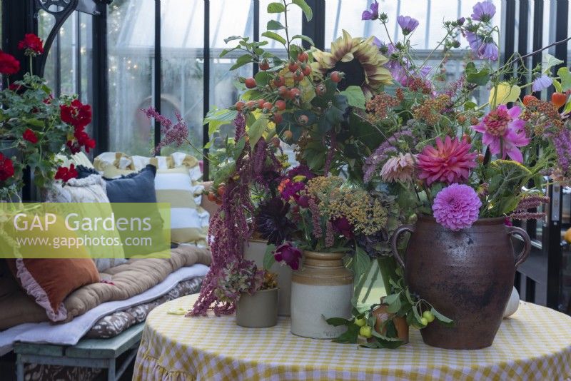 A table in the greenhouse that has a collection of vases of cut flowers, including: dahlias, sunflowers, cosmos,sedums and rose hips.