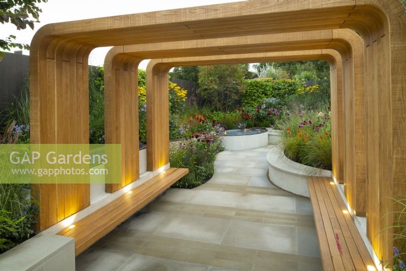 A modern timber structure with benches leading to a paved area surrounded by rills and raised herbaceous borders containing autumn planting in the Finding Our Way, An NHS Tribute Garden 