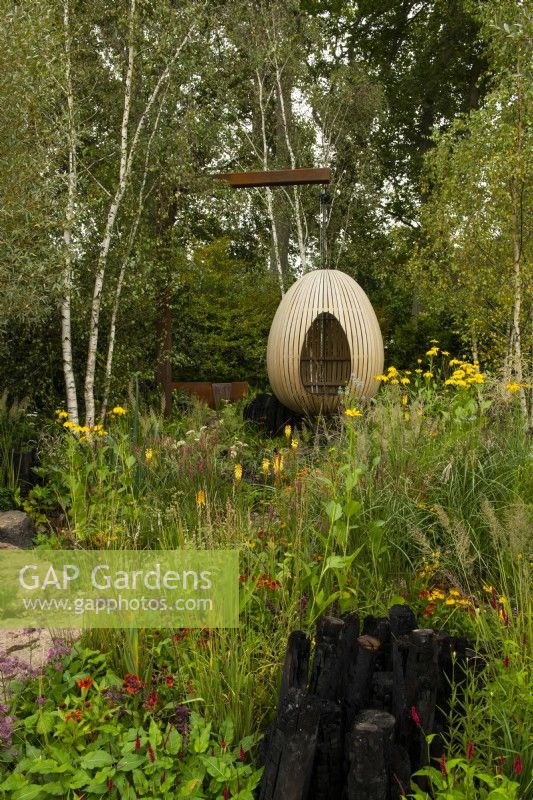 An egg shaped oak hide surrounded by woodland and herbaceous perennials bordered by charcoal logs.  
