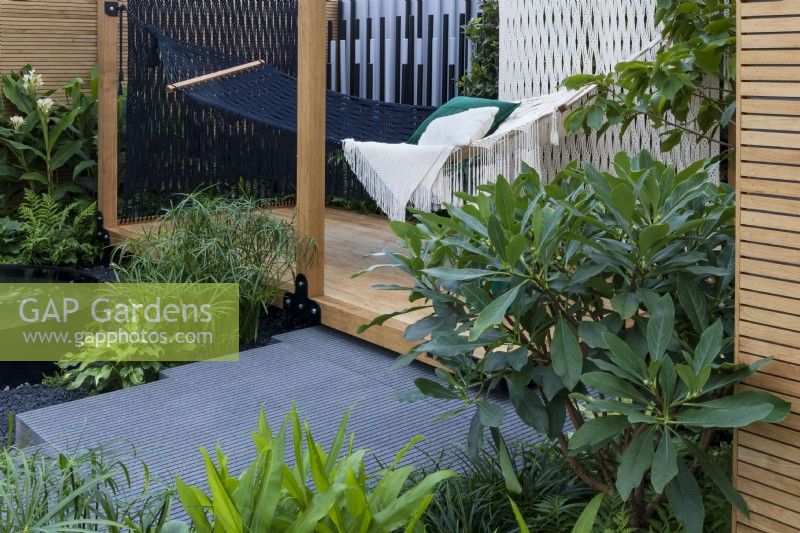 Hammock strung on wooden deck with slatted walls. Green plants in the foreground including foliage of oleander and Hedychium. Cyperus alternifolius next to water feature with white ginger lily, Hedychium coronarium behind. Calm of Bangkok.