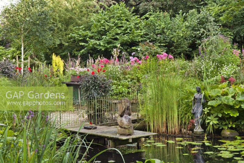 Natural pond with moisture loving plants, wooden pier and a woman statue: Darmera peltata, Dahlias in containers, water lilies, Typha laxmannii and grasses. 