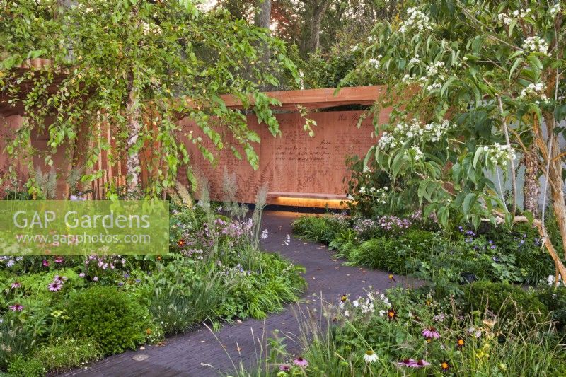 Pathway through mixed borders to the bench and wall panel. The Florence Nightingale Garden, RHS Chelsea Flower Show 2021