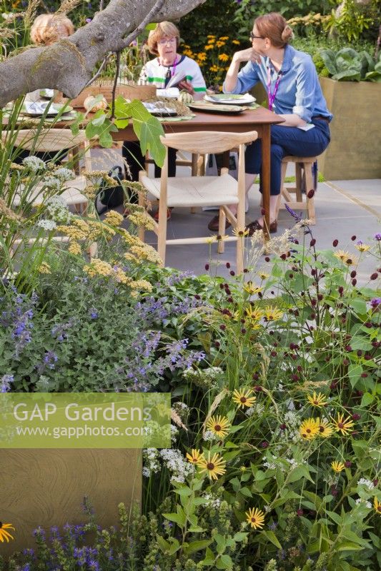 bed with sanguisorbas, rudbeckias, achilleas, catmint and salvias. People relaxing in the garden in the background. The Parsley Box Garden at Chelsea Flower Show 2021