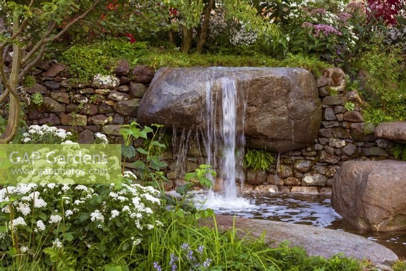 Psalm 23 Garden. Naturalistic cascade of water from granite, worn rocks in drystone wall into tranquil pool. Featuring Eupatorium 'Lucky Melody', Carex sp.,  and young shoots of Viburnum opulus .