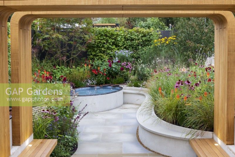 Finding Our Way: an NHS Tribute Garden. Wooden pergola leads into sunken paved courtyard space with white paving, raised pool and flower beds. Plants include Echinacea purpurea, Echinacea 'Eccentric', Dahlia 'Sam Hopkins', Pennisetum alopecuroides 'Hameln', and Kniphofia.