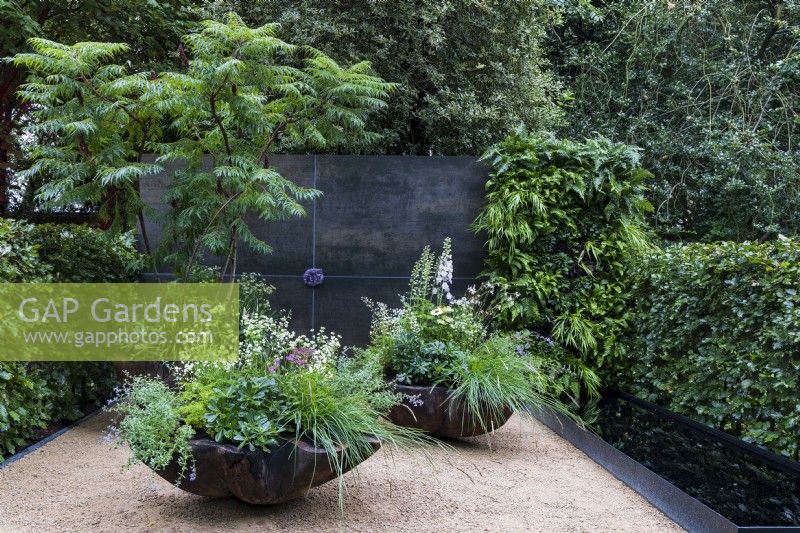 Stolen Soul Garden. Three sculptural, scalloped, wooden containers, with mainly green and white planting stand on a bare surface, with black wall behind, set with an amethyst crystal in the centre. Plants include a stag's horn sumach tree, Rhus typhina, astrantia, evergreen foliage, delphiniums, nepeta, and grasses.