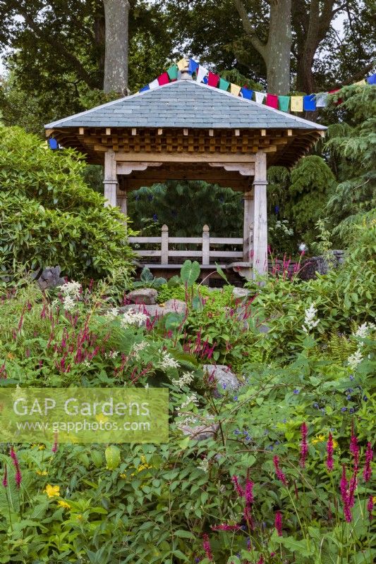 Trailfinders' 50th Anniversary Garden. Wooden shelter resembling Nepalese religious building, with prayer flags strung across. Rhododendrons flank perennial planting including Persicaria affinis 'Darjeeling Red', Persicaria amplexicaulis, bamboo Fargesia rufa, Potentilla fruticosa, and Inula.