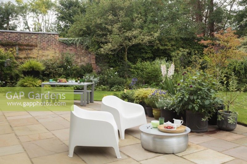 Stone patio area with metal table and modern chairs looking out onto garden with groups of pots, borders, mature shrubs and trees - Cheshire - July