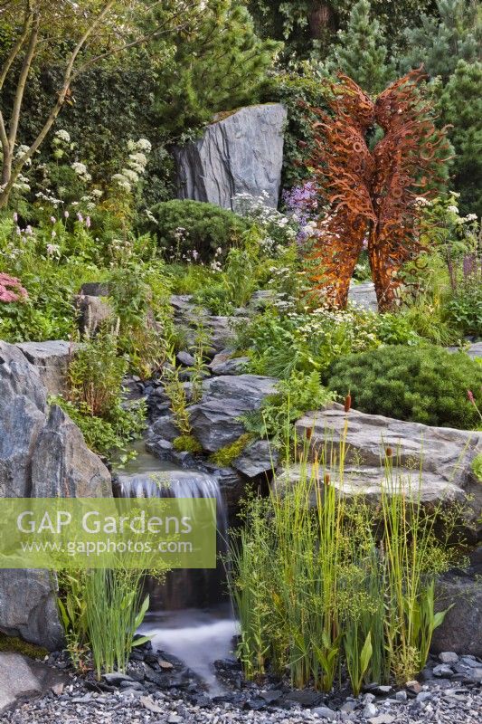 View over waterfall, with planting such as Typha laxmannii among rocks, to rusty metal sculpture. Bodmin Jail: 60Ã‚Â° East - A Garden Between Continents, RHS Chelsea Flower Show 2021