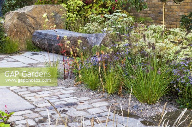 The M and G Garden. Paved area with rustic carved bench and a mix of gravel and paving for improved drainage. Autumn planting combination including  Pennisetum alopecuroides 'Cassian', Eurybia x herveyii, Sporobolus heterolepis, and Selinum wallichianum.