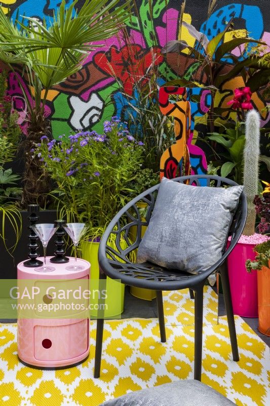 Pop Street Garden. Chair with silver cushions, cylindrical table and cocktail glasses sits on coloured patterned flooring with a street art mural by Robert Littleford behind. Plants include Aster frikartii 'Monch', Miscanthus sinensis 'Dronning Ingrid', Canna indica 'Russian Red', and hardy palm Trachycarpus fortunei.
