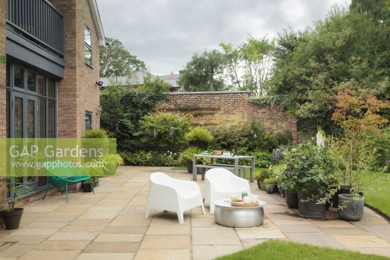 Contemporary house with stone patio area with metal table and modern chairs looking out onto garden with groups of pots, borders, mature shrubs and trees - Cheshire - August