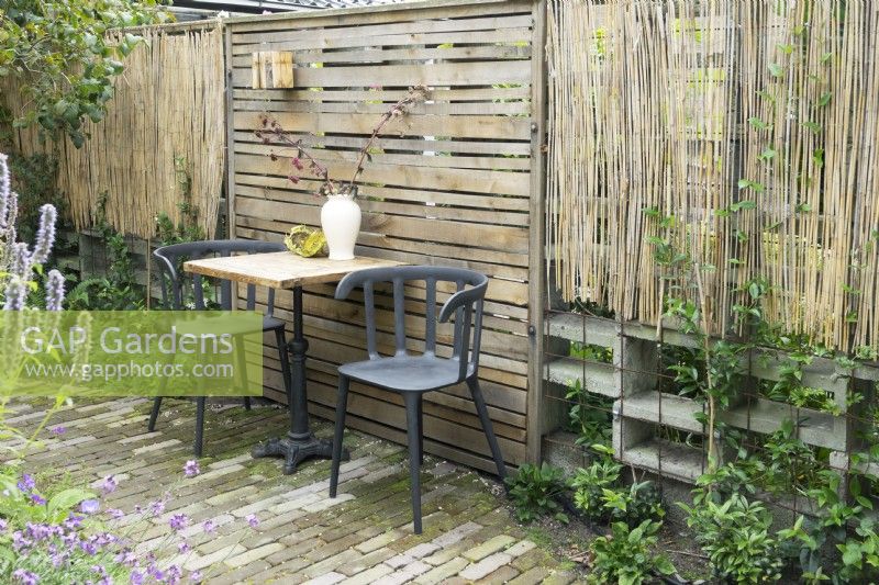 Slatted fence panel with table and chairs in front, either side a boundary of open concrete blocks with bamboo screen plus metal trellis with climbers.