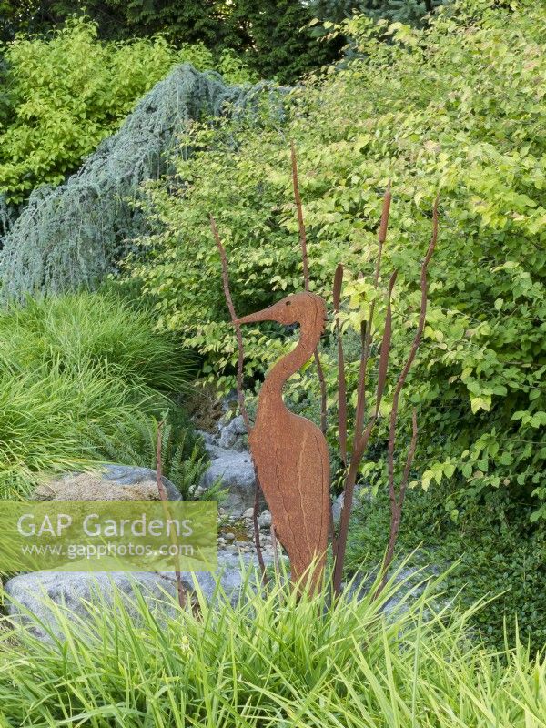Metal sculpture of heron and bullrushes in dry stream bed featuring foliage of daylilies, spike winterhazel and weeping blue atlas cedar