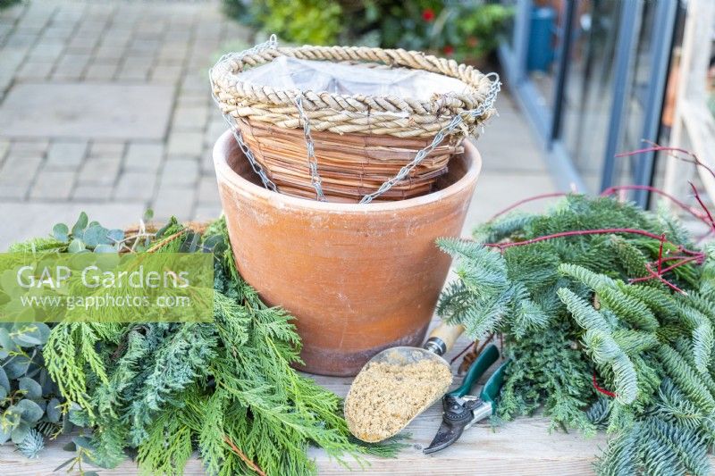 Hanging basket, compost scoop, sand, secateurs and evergreen foliage laid out on a wooden surface