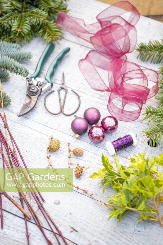 Evergreen foliage, dogwood twigs, ribbon, pine cones, baubles, wire, pruning scissors and secateurs laid out on a wooden surface