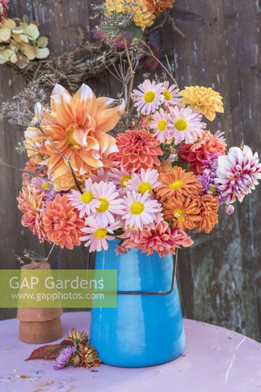 Pink and Orange flower arrangement of Dahlias and Chrysanthemums with dried fennel in blue enamel jug on table against wooden background