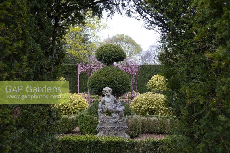 A baroque carved stone cherub statue surrouned by clipped Box hedge and Yew topiary in the sculpture garden at Thenford Gardens and Arboretum, Thenford, Banbury, Oxfordshire, UK