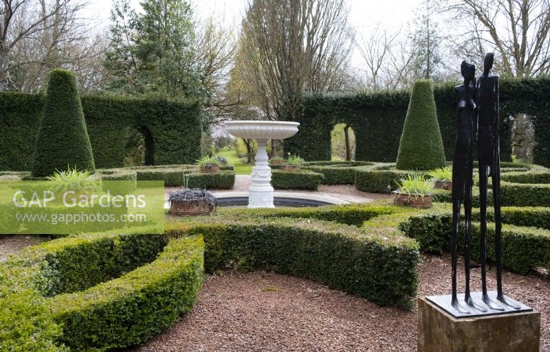 A marble urn and bronze sculpture 'The Couple' surrounded by clipped box and yew topiary in the Knot Garden at Thenford Gardens and Arboretum, Thenford, Burford, Oxfordshire, UK