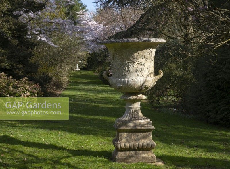 A carved stone urn and Prunus blossom on Lannings Walk at Thenford Gardens and Arboretum, Thenford, Burford, Oxfordshire, UK