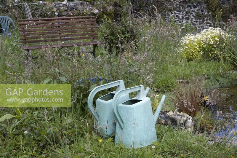 Two old plastic watering cans sit beside a pond with an old bench and chair in the background. Derrydown. NGS garden. July. Summer. 