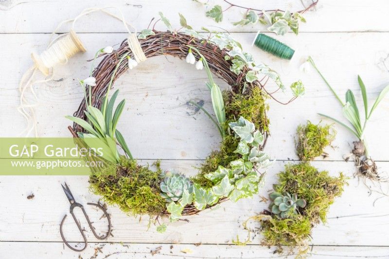 Completed wreath, moss, snowdrops, wire, string and pruning scissors laid out on a wooden surface