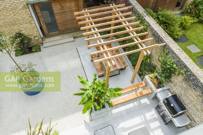 Aerial view of a contemporary courtyard garden with pergola, outdoor seating, barbeque, large planters and plants with a tropical appearance. September