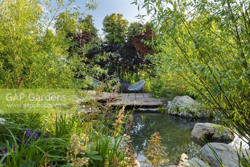 View across pond to seating area on wooden deck. RHS Garden For A Green Future. Designed by Jamie Butterworth. RHS Hampton Court Palace Garden Festival Show, July 2021