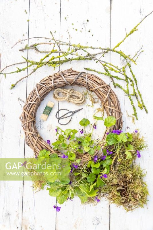 Lichen covered sticks, moss, violas, wreath, rope, wire and gardening scissors laid out on a wooden surface