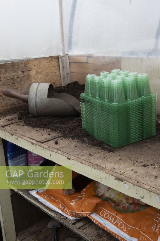 A potting bench in a small commercial nursery with wooden handled scoop, compost and a bulk lot of seed trays in the foreground and a bag of compost underneath. Spring. 