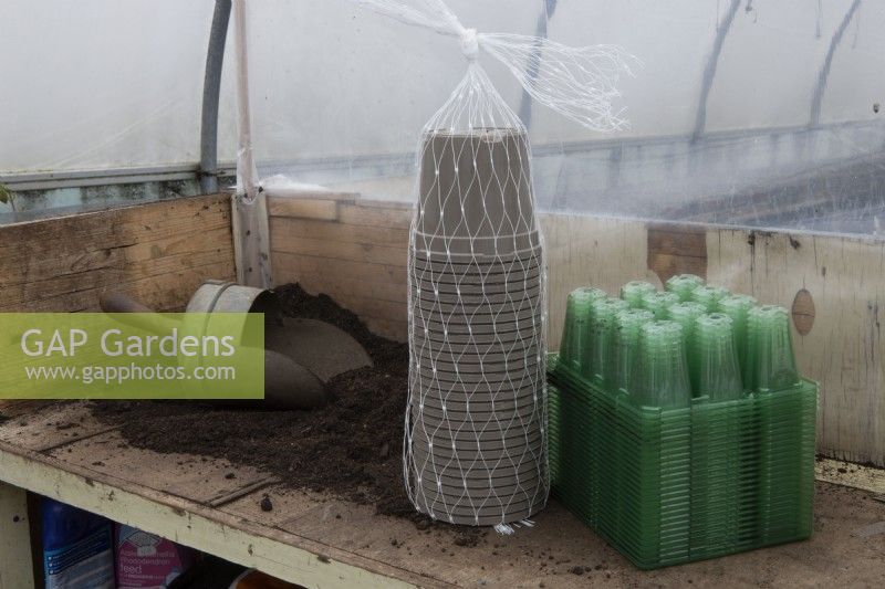 A bulk load of green plastic seed trays and grey plant pots sit on a potting bench beside a pile of compost and a wooden handled scoop in a small commercial nursery. 