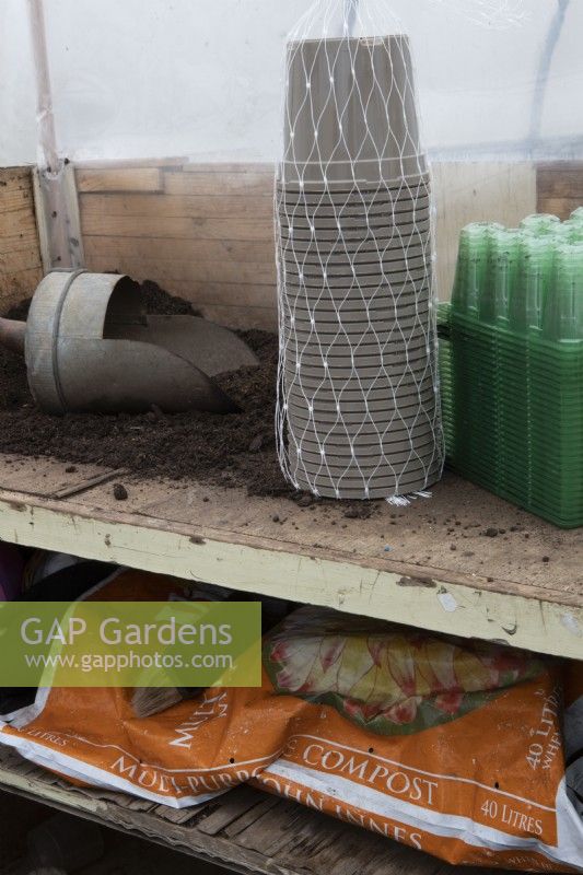 A bulk load of green plastic seed trays and grey plant pots sit on a potting bench beside a pile of compost and a wooden handled scoop in a small commercial nursery with a bag of compost below. Spring.