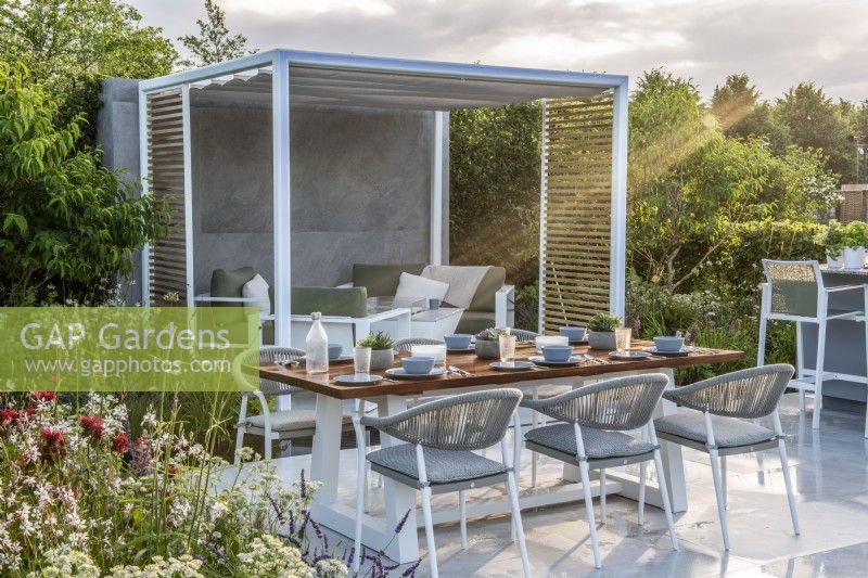 Morning sun on patio with dining table and metal pergola with seating under.  Lower Barn Farm: The Bounce Back Garden, RHS Hampton Court Palace Garden Festival 2021