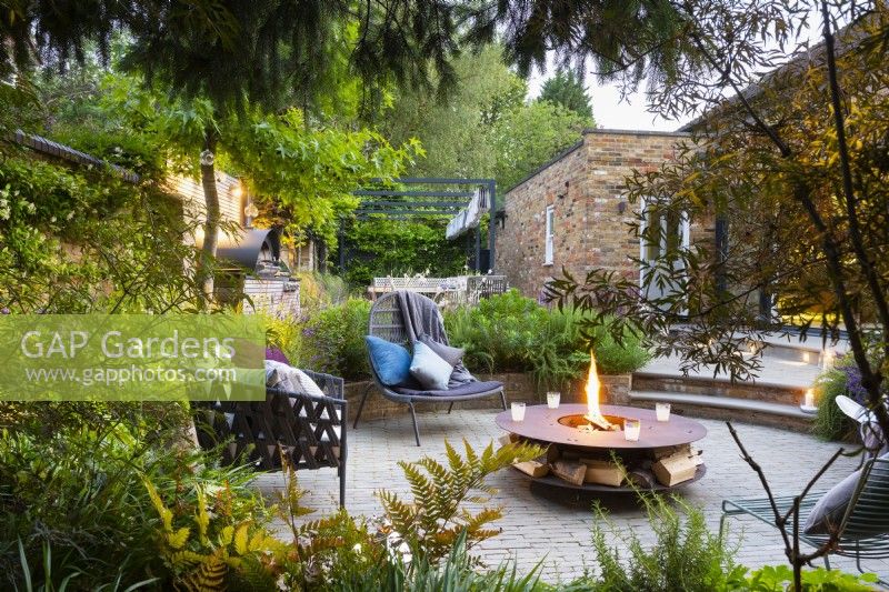 View across beds with shade tolerant plants to sunken seating area with freestanding chairs and fire pit.