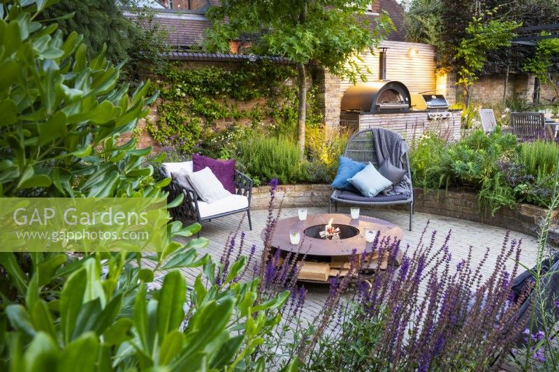 View across flower beds to sunken seating area with freestanding chairs and fire pit.